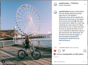 An Instagram image capturing an image of a brown-haired woman standing with a bike on a pier with a large ferris wheel and harbor water in the background on a sunny day, accompanied by a caption.