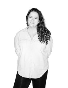 A black and white headshot of a woman with shoulder-length brown hair standing and posing with her arms behind her back.